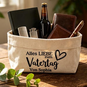 Puede incluir: Una cesta de tela beige llena de un cuaderno negro, dos encendedores plateados, una botella de cerveza oscura, dos carteras de cuero marrón y un bolígrafo. La cesta tiene texto negro que dice "Alles Liebe zum Vatertag Von Sophia" con un corazón.
