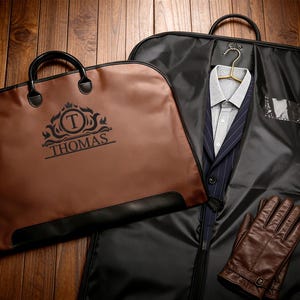 Può includere: Una borsa portabiti marrone con il nome "THOMAS" stampato sopra, accanto a una borsa portabiti nera contenente un abito e un paio di guanti in pelle marrone. L'abito è blu navy con gessati bianchi e una camicia grigio chiaro.