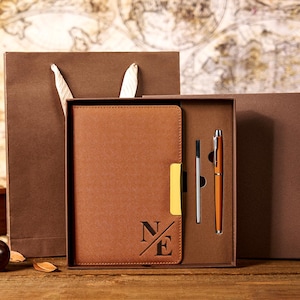 May include: A brown leather journal and pen set, embossed with the initials "N/E" on the cover. The set includes a matching pen and refill, presented in a brown gift box with a brown paper gift bag.