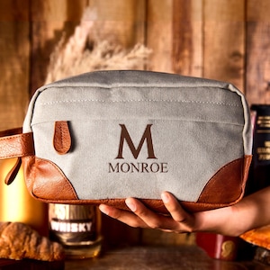 Puede incluir: Neceser de lona gris con detalles en cuero marrón y un asa. La bolsa presenta la inicial "M" y el nombre "Monroe" en marrón. Ideal para guardar artículos de aseo y personales.