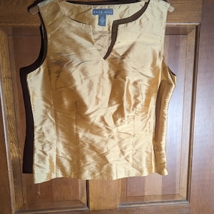 May include: Gold-colored sleeveless top with a V-neckline. The top is made of a shiny, textured fabric and is hanging on a wooden hanger. The tag reads "KATE HILL".
