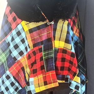 Può includere: Uno scialle di plaid colorato con un colletto in pelliccia sintetica nera. Lo scialle è fatto di un patchwork di diversi tessuti a quadri in rosso, blu, giallo e verde.
