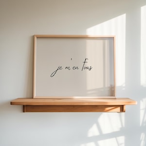 Puede incluir: Un cuadro enmarcado con la frase "je m'en fous" en elegante escritura negra. La obra de arte se muestra sobre un fondo beige claro, dentro de un marco de madera natural. El marco descansa sobre una estantería de madera contra una pared blanca.