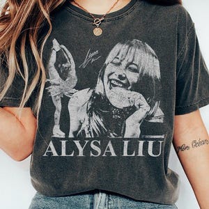 May include: Dark gray t-shirt featuring a black and white image of Alysa Liu, a figure skater, with her name printed below. The graphic includes a picture of Alysa Liu skating and a signature.