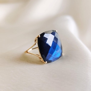 May include: A gold ring featuring a large, square-cut, faceted dark blue gemstone. The stone has a cushion-like shape with multiple reflective surfaces, held in place by a thin, delicate gold band that splits into two prongs at the setting.