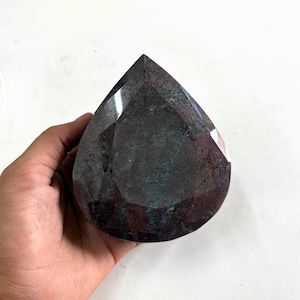 May include: A teardrop-shaped gemstone with a faceted surface, displaying a deep, mottled colour palette of dark burgundy, maroon, and forest green tones. The stone features multiple angular cuts that create reflective planes across its surface.
