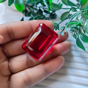 Peut inclure: Une grosse pierre précieuse rectangulaire à facettes, d'un rouge profond. La gemme est tenue dans une main, avec un feuillage vert en arrière-plan. La taille de la pierre permet à la lumière de se refléter, créant un éclat brillant. Cette pierre précieuse pourrait être utilisée en joaillerie.