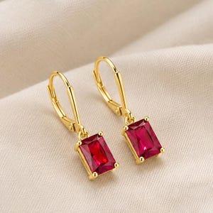 May include: A pair of gold leverback earrings featuring rectangular, faceted, deep red gemstones. The earrings have a polished gold finish and a secure hinged closure mechanism, showcasing a classic drop design with vibrant red stones.