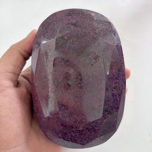 May include: A large, oval-shaped, faceted gemstone with a deep magenta and dark purple color palette. The stone features a polished surface with multiple geometric facets that reflect light, showcasing a textured, mottled appearance throughout.
