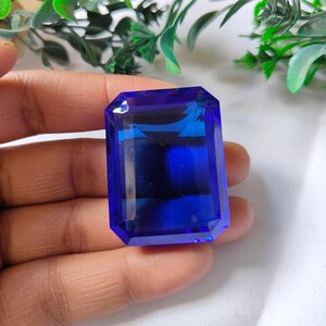 May include: A large, rectangular, faceted gemstone in a deep blue colour. The gem is held in a hand, highlighting its clarity and brilliance. The cut is an emerald cut, with a polished surface. The gem is likely a sapphire.