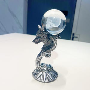 Puede incluir: Una escultura decorativa de color plateado que presenta una cabeza de lobo que sostiene un globo de cristal transparente. Dentro del globo hay un grabado detallado de una calavera. La base tiene un diseño floral.