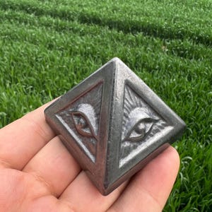 Può includere: Una piramide metallica quadrata con disegni di occhi incisi su ogni lato. La piramide è tenuta in mano su uno sfondo di erba verde. Il metallo ha un aspetto grigio e usurato.