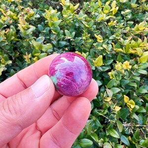 May include: A polished, spherical gemstone with a vibrant magenta hue, veined with lighter pink and a touch of green. The gemstone is held in a hand, with a backdrop of green and yellow foliage.