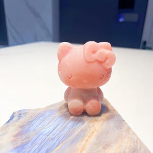 Könnte beinhalten: Eine rosa Cartoon-Katzenfigur mit Schleife, die auf einer mehrfarbigen, strukturierten Oberfläche steht. Die Katze hat einen runden Kopf, kleine Augen und eine Schleife. Die Figur besteht aus einem festen, glatten Material.