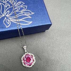May include: A silver necklace with a large, round pendant. The pendant features a central pink gemstone surrounded by a halo of smaller, clear stones. The necklace is displayed on a gray surface next to a blue box with a floral design.