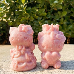 Könnte beinhalten: Zwei rosa Edelsteinfiguren einer Katzenfigur, eine mit Kleid und die andere mit einem Herz. Die Figuren haben runde Augen, eine Schleife und sind aus einem hellrosa Stein geschnitzt. Der Hintergrund ist verschwommenes grünes Laub.