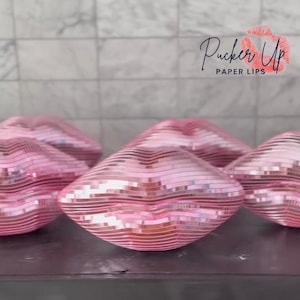 Puede incluir: Cuatro decoraciones de fiesta en forma de labios rosas con un acabado espejado. Las decoraciones están hechas de papel y tienen el texto "Pucker Up Paper Lips" sobre un fondo blanco.