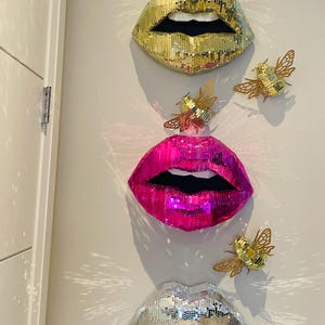 May include: Three mirrored lips, one gold, one pink, and one silver, are displayed on a white wall. Each lip is made of small, individual mirrored tiles. Three gold bee decorations are also displayed on the wall.