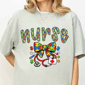 May include: A light green t-shirt with the word "NURSE" in colorful puzzle piece letters. Below the word is a bow tie made of puzzle pieces, a red stethoscope, and a medical symbol.