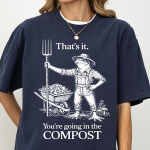 May include: Navy blue t-shirt with a graphic of a frog farmer holding a pitchfork and a wheelbarrow. The text on the shirt reads "That's it. You're going in the COMPOST."