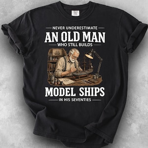 May include: A black t-shirt featuring a graphic of an older person with a beard working on a model ship at a desk with a lamp. The text reads: NEVER UNDERESTIMATE AN OLD MAN WHO STILL BUILDS MODEL SHIPS IN HIS SEVENTIES. The design is centered.