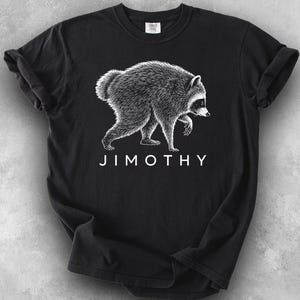 Pode incluir: Uma t-shirt preta de manga curta com uma ilustração gráfica branca de um guaxinim de perfil. Abaixo do guaxinim, o texto JIMOTHY está impresso em letras maiúsculas brancas. A t-shirt tem um decote redondo e um design padrão.