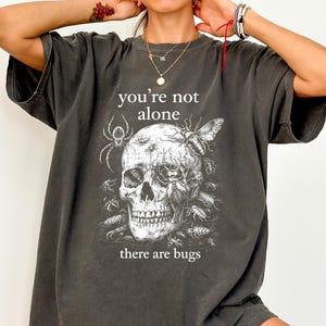May include: Dark gray t-shirt featuring a skull design with insects and the text "you're not alone there are bugs" in white. The shirt has short sleeves and a relaxed fit, suitable for casual wear.