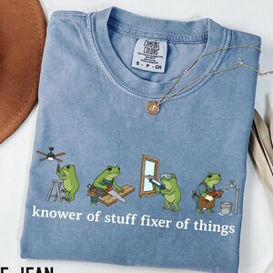 May include: Blue jean colored t-shirt with a crew neck and short sleeves. The shirt features a graphic of cartoon frogs performing home repair tasks, with the text "knower of stuff fixer of things". A gold necklace is draped across the shirt.