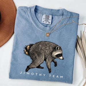 Pode incluir: Uma t-shirt de gola redonda azul-clara com uma ilustração detalhada de um guaxinim de perfil. O texto "JIMOTHY TEAM" está impresso a branco por baixo do guaxinim. A t-shirt tem uma etiqueta da marca Comfort Colors no pescoço com o tamanho S.