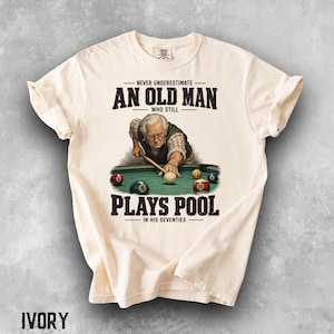 May include: An ivory t-shirt featuring a graphic of an older person playing pool with the text NEVER UNDERESTIMATE AN OLD MAN WHO STILL PLAYS POOL IN HIS SEVENTIES. The illustration shows a person in a vest aiming a cue stick at billiard balls.