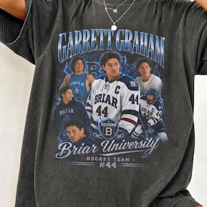 Könnte beinhalten: Dunkelgraues T-Shirt mit einem Grafikdesign. Das Design enthält den Text "GARRETT GRAHAM", "Briar University Hockey Team #44" und Bilder eines Hockeyspielers. Das Wort "BRIAR" ist ebenfalls enthalten.
