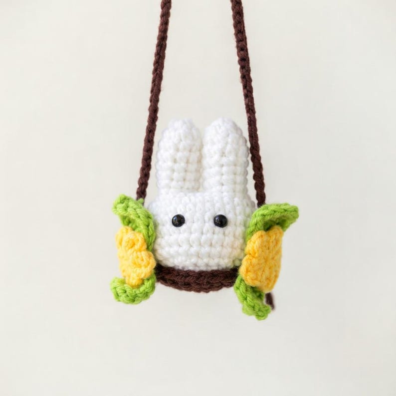 May include: A white crocheted rabbit head with black bead eyes, nestled in a brown base. The rabbit is flanked by two yellow flowers with green leaves on either side. Two long brown crocheted straps extend upward from the base of the rabbit.