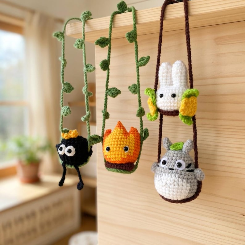 May include: Three crocheted hanging ornaments featuring a black soot sprite with a gold star, an orange and yellow flame character, and a white and gray creature with green leaves, all suspended by green vine or brown yarn ropes.