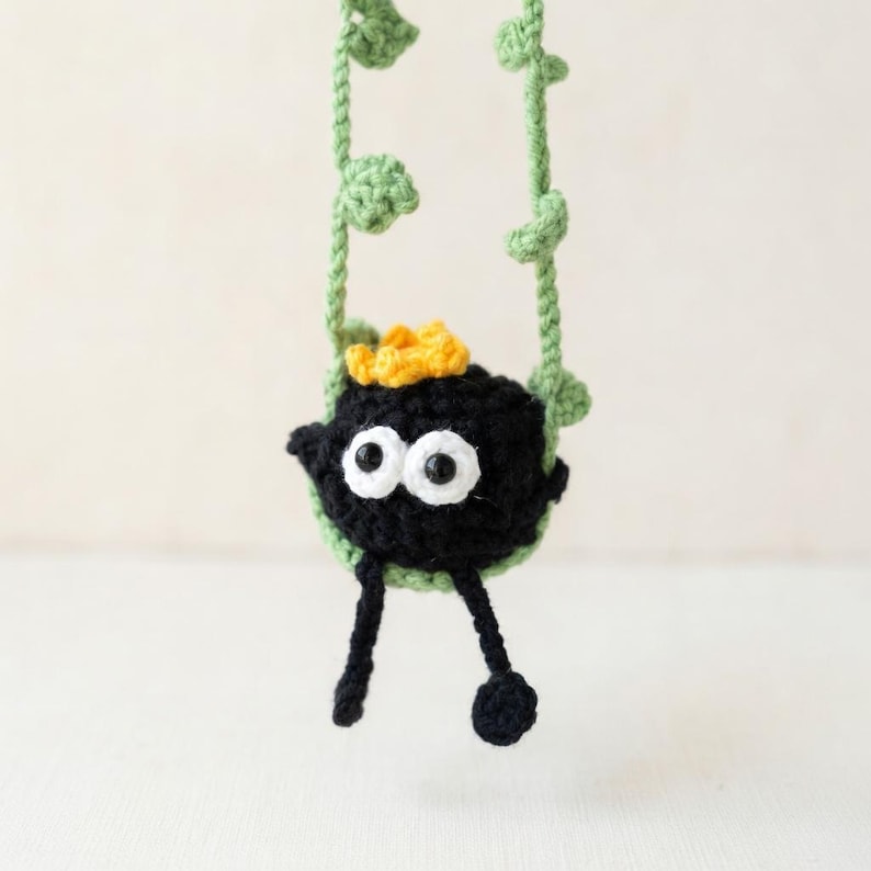 May include: A small, round, black crocheted creature with two large white eyes, a yellow tuft on top, and two dangling black legs. It is suspended by two light green crocheted vine-like strands that feature small, textured leaf-shaped accents.