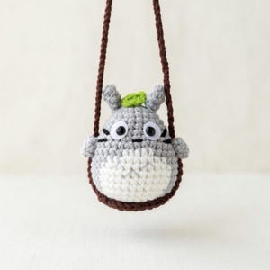 May include: A gray and white crocheted creature ornament with a small green leaf on its head, black plastic eyes, and dark brown stitched whiskers. The figure is suspended by a dark brown braided cord, creating a hanging decorative accessory.