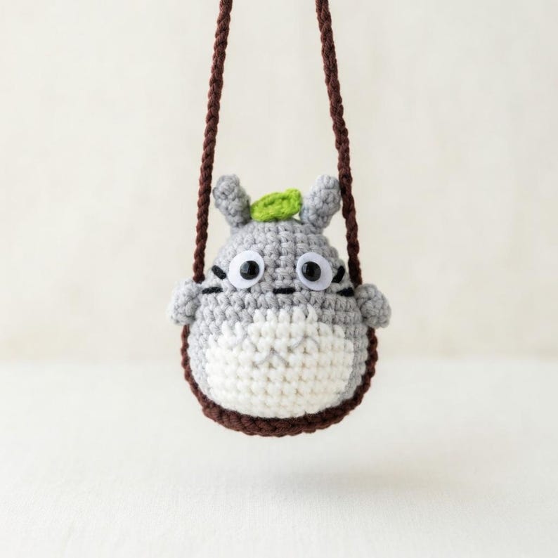 May include: A gray and white crocheted creature ornament with a small green leaf on its head, black plastic eyes, and dark brown stitched whiskers. The figure is suspended by a dark brown braided cord, creating a hanging decorative accessory.