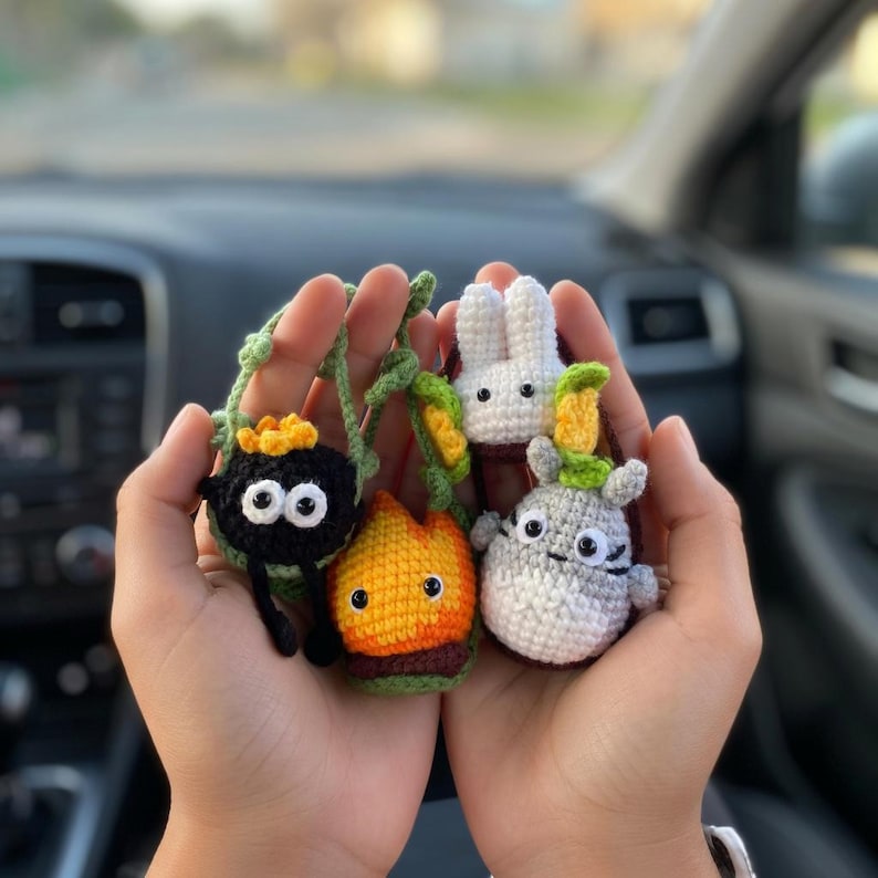 May include: A collection of four small crocheted figures including a black soot sprite with a yellow star, a white rabbit, a flame-shaped character in orange and yellow, and a grey and white creature with small ears and a green leaf on its head.