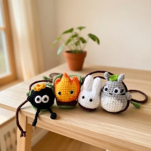 May include: Four small, handmade crochet pouches with long brown straps. The characters include a black soot sprite with a yellow star, a flame-shaped orange and yellow figure, a small white rabbit-like creature, and a grey round creature.