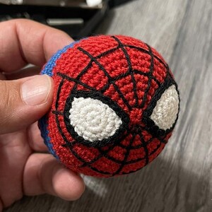 以下が含まれることがあります： スパイダーマンのマスクのデザインが施された手作りの編みぐるみボール。ボールは主に赤色で、黒いクモの巣、白い目のディテール、青色のアクセントがあります。手作りのアイテムが手に持たれています。