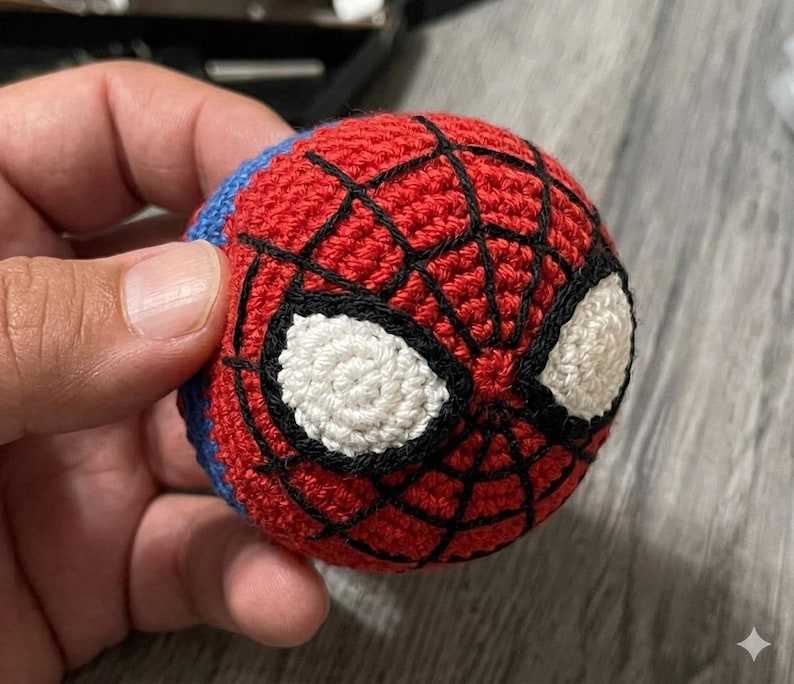 以下が含まれることがあります： スパイダーマンのマスクのデザインが施された手作りの編みぐるみボール。ボールは主に赤色で、黒いクモの巣、白い目のディテール、青色のアクセントがあります。手作りのアイテムが手に持たれています。