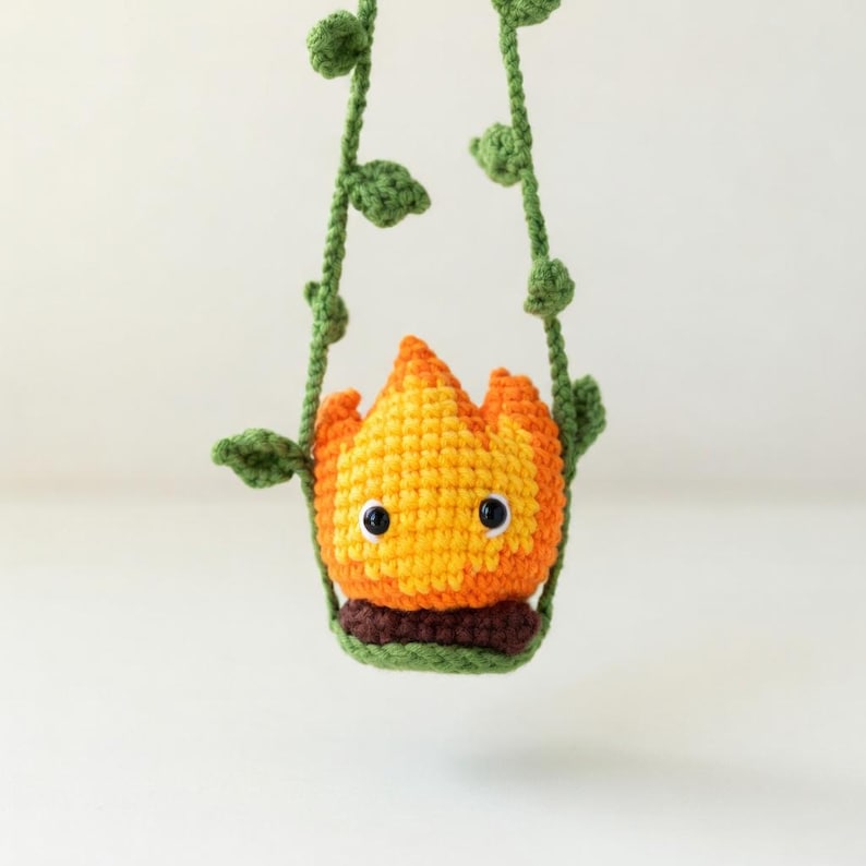May include: A crocheted orange and yellow flame character with black safety eyes, sitting in a dark brown base. The figure is suspended by two green crocheted vine-like strands featuring small green leaves, creating a hanging display.