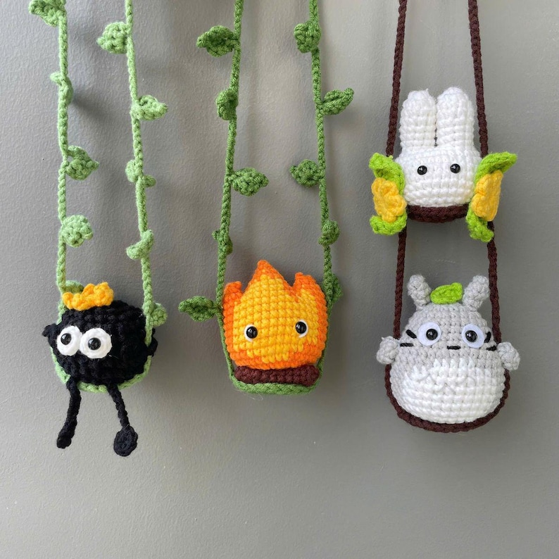 May include: Three crocheted hanging ornaments featuring a black soot sprite with yellow crown, an orange and yellow flame character, and a white rabbit-like creature above a gray and white round character, all suspended by green or brown yarn vines.