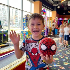 以下が含まれることがあります： 赤いと黒の編みぐるみのスパイダーマンボールを、白い目のディテールで持っている笑顔の少年。スパイダーマンのグラフィックTシャツを着ています。背景には、さまざまなゲーム機を備えたアーケードがあります。