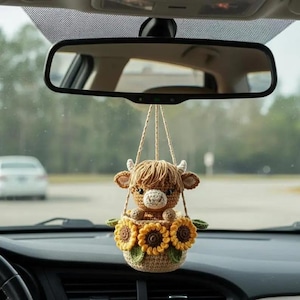 May include: A crocheted car charm featuring a brown Highland cow with a cream-colored face, small horns, and a shaggy mane. The cow sits in a basket adorned with yellow sunflowers and green leaves, suspended by beige strings from a car's rearview mirror.
