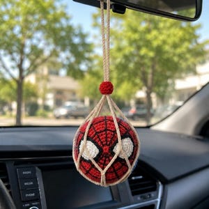 以下が含まれることがあります： 赤、黒、白の編み物スパイダーマンカーチャームが車のバックミラーから吊り下げられています。チャームはクモの巣のデザインと白い目のディテールを持つ球体です。ベージュの紐で吊り下げられています。