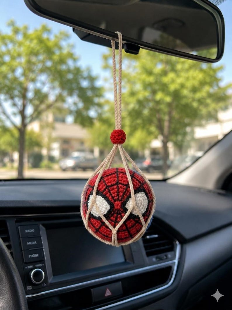 以下が含まれることがあります： 赤、黒、白の編み物スパイダーマンカーチャームが車のバックミラーから吊り下げられています。チャームはクモの巣のデザインと白い目のディテールを持つ球体です。ベージュの紐で吊り下げられています。