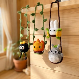 May include: Three crocheted hanging ornaments featuring a black soot sprite with a gold star, an orange flame character, and a white rabbit-like creature above a grey round character, all suspended by green vine-like or brown yarn cords.