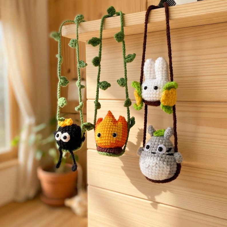 May include: Three crocheted hanging ornaments featuring a black soot sprite with a gold star, an orange flame character, and a white rabbit-like creature above a grey round character, all suspended by green vine-like or brown yarn cords.