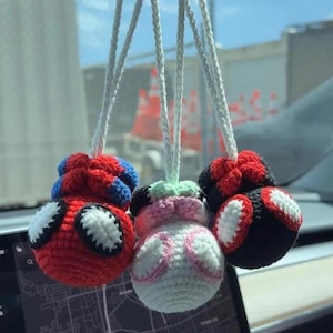 May include: Three handmade crochet car charms. One is red with blue and white accents, another is white with pink and green accents, and the last is black with red and white accents. Each charm hangs from a white string.