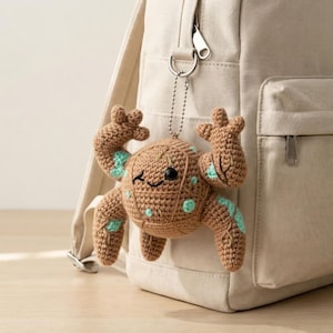 May include: A crocheted brown crab keychain with teal accents and a smiling face is attached to a beige backpack. The crab has a round body, six legs, and two claws. The keychain is made of yarn and has a metal chain.